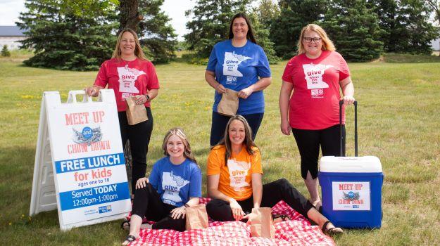 Women in red, blue, and orange Give Where You Live shirts hold bang lunches in park next to Meet Up and Chow Down sandwich board and cooler