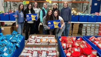 Volunteers hold "United is the Way" signs surrounded by boxes of non-perishable food soon to be packed into Buddy Backpacks at UWNEMN's Chisholm warehouse.