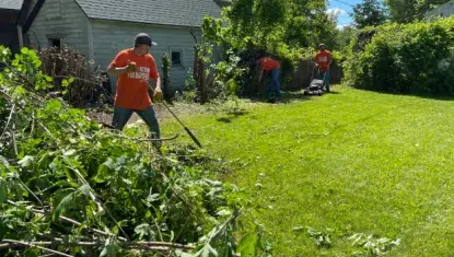 2024 Day of Action volunteers in orange t-shirts rake and mow a Keewatin residents' lawn