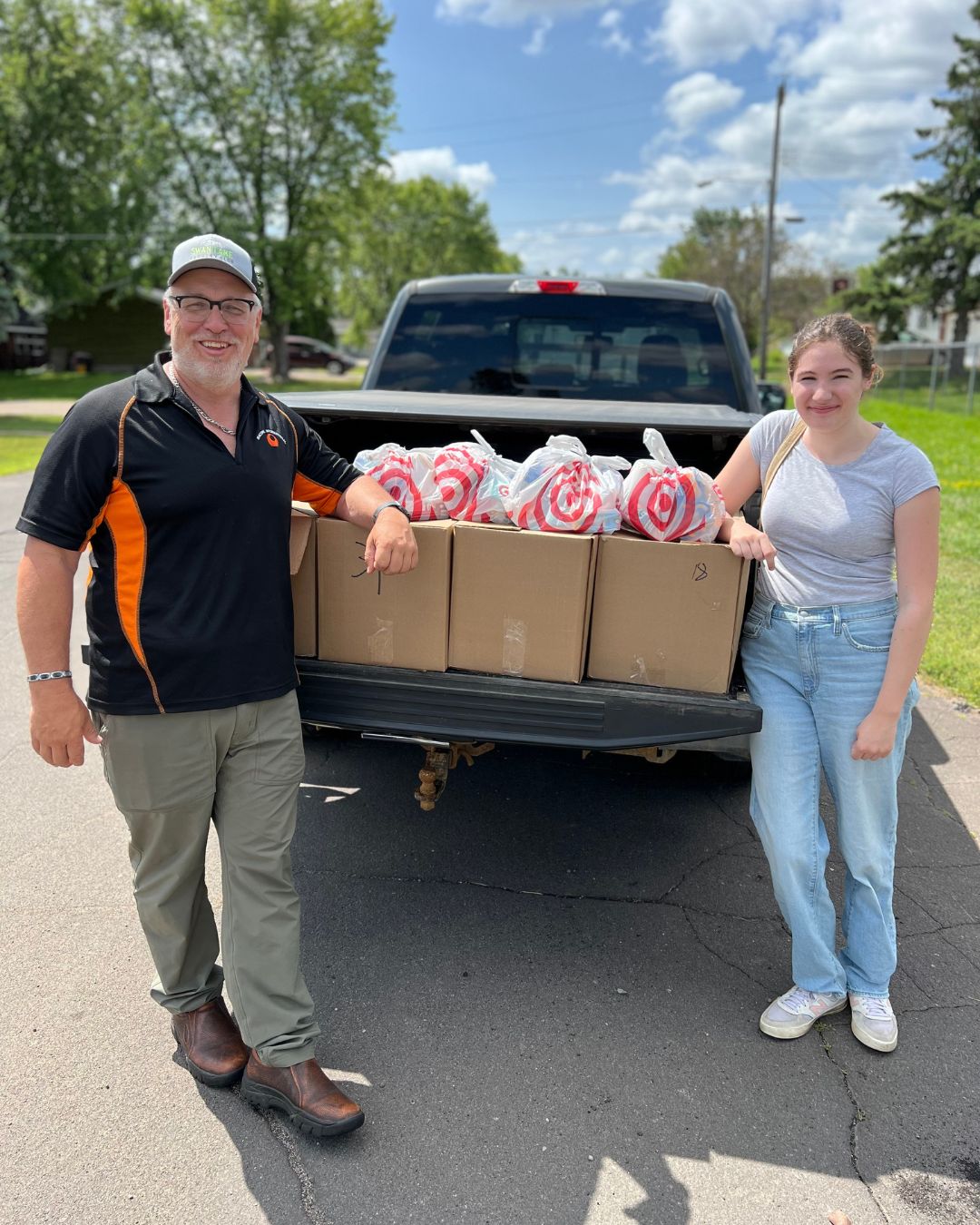 Volunteers lean on the tailgate of a truck, a male on the left and female on the right. In the tailgate are boxes filled with "Summer Buddy Backpacks."